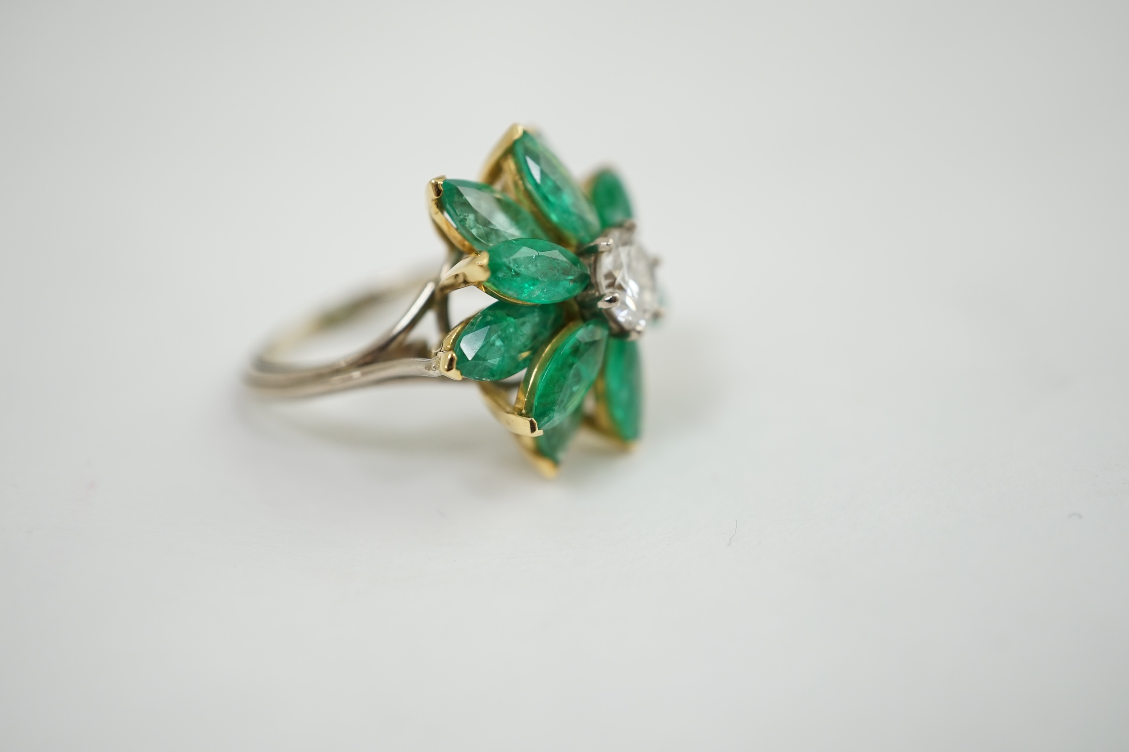 An 18ct white gold and yellow gold, emerald and diamond flowerhead dress ring, the central circular cut diamond surrounded by eleven navette shaped emerald 'petals', all claw set